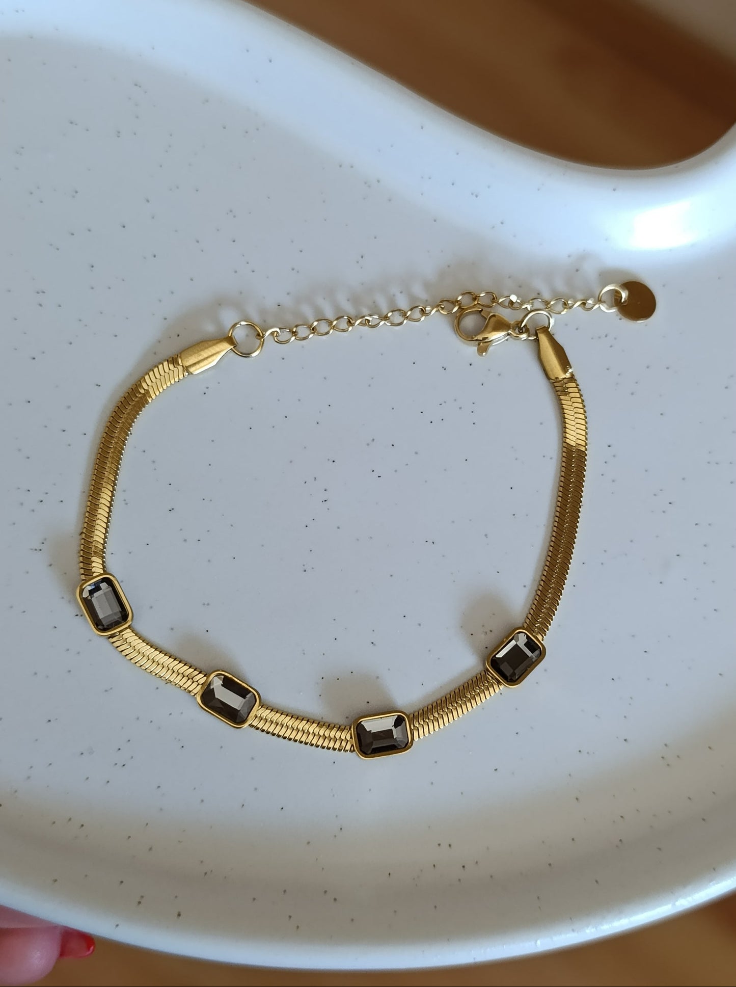 Χρυσό βραχιόλι stainless steel τύπου snake με σκούρες ορθογώνιες πέτρες, αλυσίδα επέκτασης και lobster κούμπωμα πάνω σε λευκή κεραμική πιατέλα.