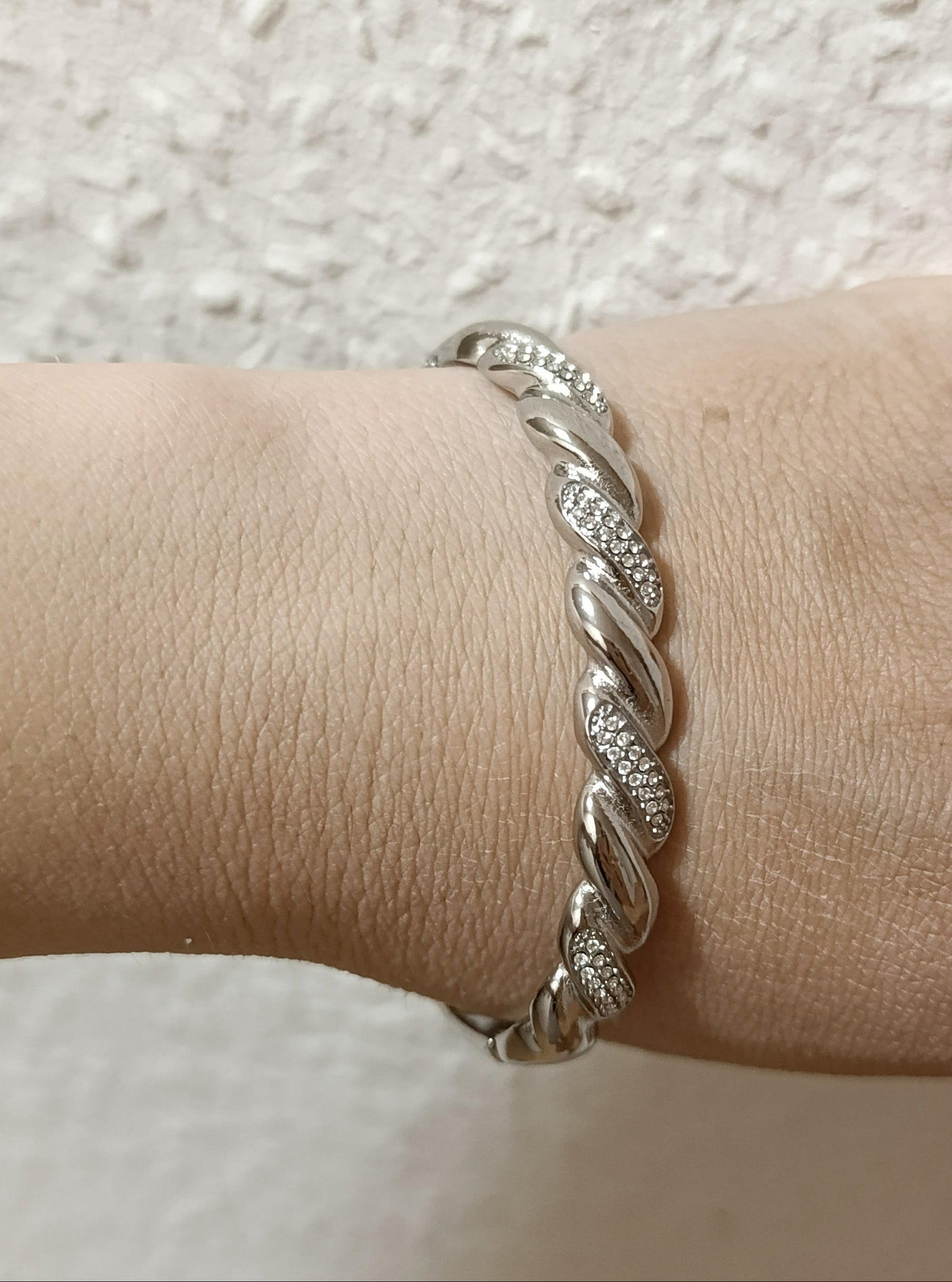 Ασημί χειροπέδα από stainless steel φορεμένη στον καρπό, με στριφτό πλεκτό σχέδιο και λαμπερές λεπτομέρειες ζιργκόν.